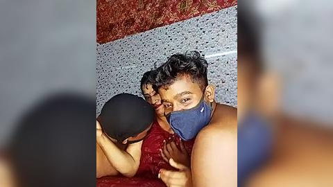 Media: Video of a young South Asian man with short, curly hair and a blue face mask, wearing a red tank top, kissing a young woman with long, wavy hair, also in a red tank top. They are in a bathroom with a tiled wall and a speckled floor.