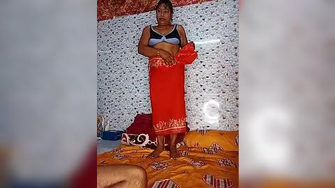 Media: Video of a middle-aged woman in a blue bra and red sarong, standing in a tiled room with a white and red-patterned wall.