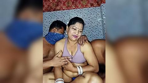 Media: Video of a South Asian woman with light brown skin, dark hair, and a medium build, wearing a lavender bra, lying on a bed with a man, partially visible, behind her. The background features a red and blue patterned cloth.