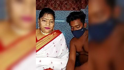 Media: Video of an elderly Indian woman in a white sari with red and yellow patterns, and a young man with a mask, standing indoors with a blue wall and red patterned fabric in the background.