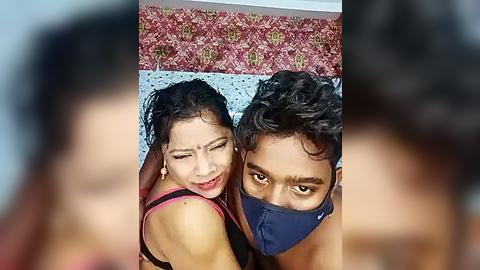 Media: A video of two young South Asian individuals, a man and a woman, smiling and wearing masks. They are in a colorful, patterned room with a floral wallpaper.