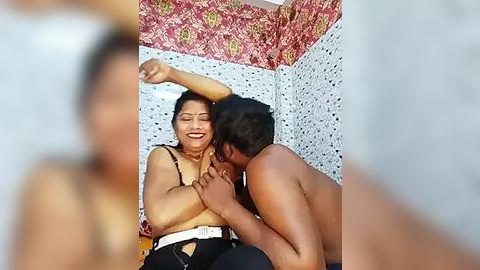 Media: A video of a South Asian woman with medium skin tone, dark hair, and a bright smile, wearing a white top and black pants, raising her arm in a corner with floral wallpaper, while a shirtless South Asian man with dark hair kisses her shoulder.