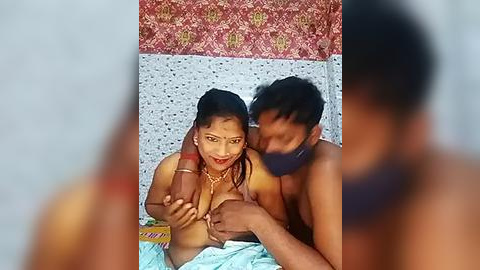 Media: Video: A topless South Asian woman with medium skin tone and dark hair, wearing a necklace, breastfeeding a shirtless man with dark skin. Background features a patterned wall and a blurred person.