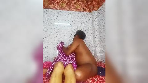 Media: A video shows a nude, dark-skinned man embracing a naked woman with a pink patterned dress, both on a red bed with white mosaic-tiled walls. The image is blurry.