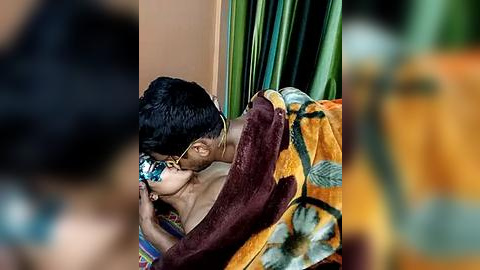 Media: Video of a South Asian couple passionately kissing in a dimly lit room. The man, with dark hair, wears sunglasses, and the woman, with long dark hair, is wrapped in a colorful blanket. Green curtains are in the background.