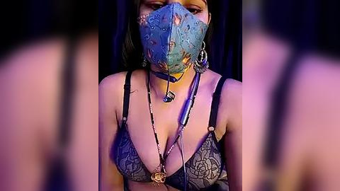 Media: Video of a woman with medium skin tone and dark hair, wearing a blue lace mask, revealing her cleavage in a gray lace bra, with a long necklace, in a dimly lit room.