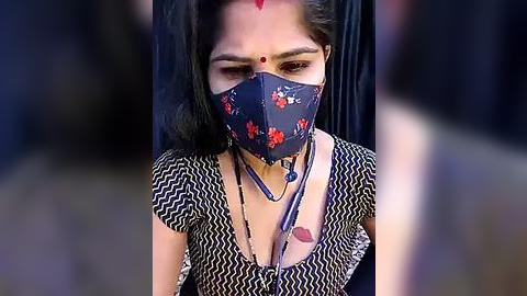 Media: Video of a South Asian woman with medium skin tone, wearing a navy blue floral face mask, traditional white and black chevron-patterned blouse, and red dot on forehead, set against a blurred background.