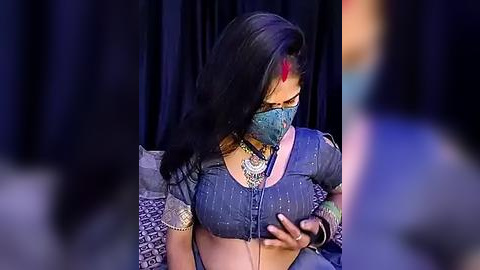 Media: Video of a woman with long black hair, wearing a blue saree with intricate embroidery, a mask, and red bindi. She holds a phone, seated against a dark, patterned background.