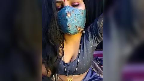 Media: Video of a South Asian woman with long black hair, wearing a blue mask and a low-cut blouse, sitting with arms raised, indoors.