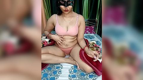 Media: Video of a curvy, fair-skinned woman in pink lingerie, sitting cross-legged on a bed with red and blue floral bedding, wearing a black mask, holding a red purse, with blurred figures in the background.