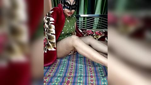 Media: Video of a woman in a green dress, black mask, and red robe with tiger print, sitting on a bed with patterned blanket, surrounded by green curtains.