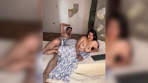 Media: A video of a naked man and woman lying on a bed, covered by a blue plaid blanket. They are in a dimly lit room with beige walls and a wooden floor.