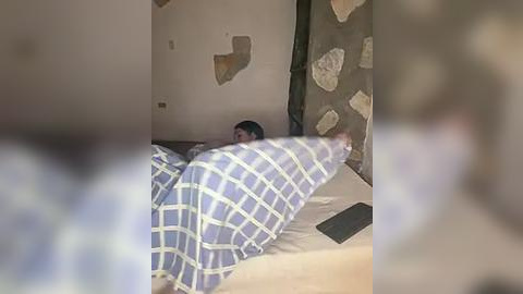 Media: A video shows a man in a bed, covered by a blue-and-white checkered blanket, with a wall behind him featuring a large, uneven patch of plaster. The scene appears dimly lit.