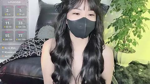 Media: Video of an Asian woman with long, wavy black hair, wearing a black face mask and a black top. She sits on a black leather couch in a room with a green plant and a yellow wall.