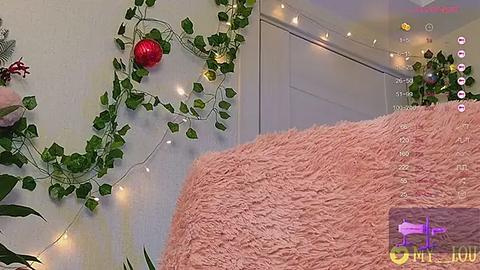 Media: A video of a cozy bedroom corner with a fluffy pink rug, a white wall adorned with green ivy and red baubles, and string lights.