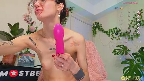 Media: Video of a topless, slender, light-skinned woman with glasses, wearing a pink dildo, in a cozy room with plants, calendar, and a 'MostBee' watermark.