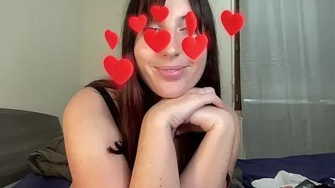 Media: Video of a young woman with long, straight brown hair, fair skin, and a small nose ring, smiling with red heart stickers over her eyes. She's in a bedroom, leaning on her hands, wearing a black tank top.