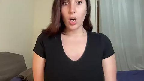 Media: A video of a young woman with medium skin tone, wearing a black V-neck t-shirt, and long dark hair. She has a surprised expression, mouth open, and is standing indoors against a beige wall and blue curtains.