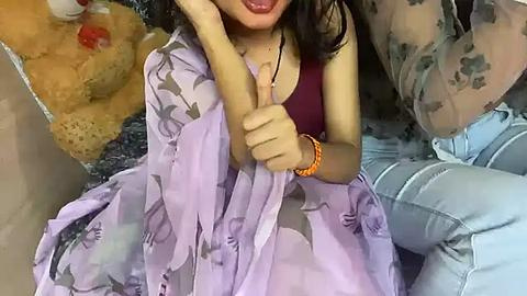 Media: Video of an Asian woman with long black hair, wearing a purple floral dress and blue jeans, giving a thumbs-up, seated on a yellow cushioned chair.