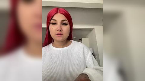 Media: Video of a young woman with long, bright red hair, wearing a white ribbed sweater, standing in a modern, minimalist room with white walls and ceiling.