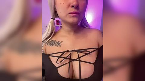 Media: Video of a fair-skinned woman with platinum blonde hair, wearing a black off-shoulder top with a laced front, showcasing ample cleavage. She has a floral tattoo on her right shoulder. Background is blurry, with purple and white lighting.