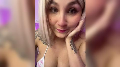 Media: Video of a young, light-skinned woman with platinum blonde hair, medium-sized breasts, and multiple tattoos on her arms, wearing a white and pink bra. She has a nose piercing and a playful smile. Background is blurred.