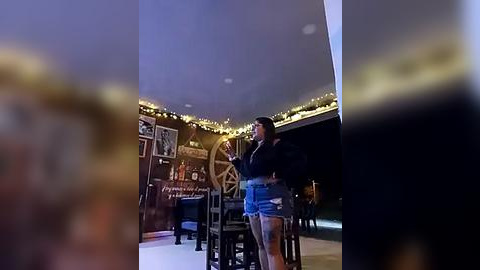 Media: Video of a woman singing on a stage with a wooden backdrop featuring a large ship's wheel and string lights. She wears a black jacket and denim shorts. Background is blurred.