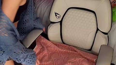 Media: A video of a person with light skin, wearing a blue lace sweater, holding a white and black patterned backpack. The background shows a pink quilted blanket and a gray pillow.