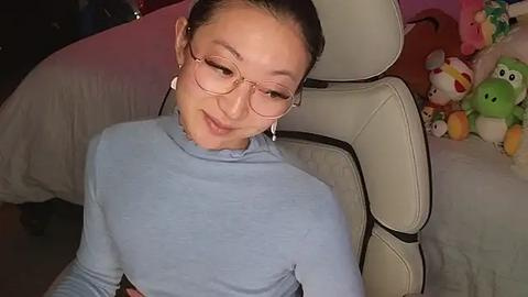 Media: Video of an Asian woman with fair skin, wearing glasses, a light blue turtleneck, and white earbuds, sitting on a white leather chair. Background features plush toys, including a green frog and a pink bunny, on a bed with white sheets.