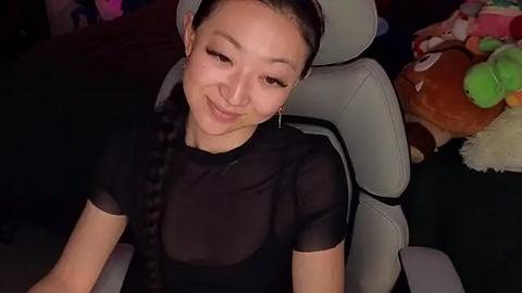 Media: Video of a young Asian woman with fair skin, black hair tied back, wearing a sheer black top, smiling. She is seated in a modern grey gaming chair, surrounded by plush toys and colorful stuffed animals in a dimly lit room.