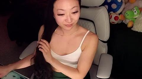 Media: Video of an Asian woman with long black hair, wearing a white tank top and green pants, sitting on a white gaming chair. Background features stuffed toys and a blue polka-dot cushion.