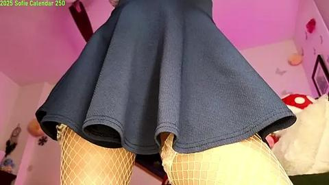 Media: Video of a woman's lower body in a short, pleated blue skirt, yellow fishnet stockings, and a white plush toy in a pink-themed room.