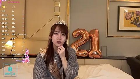 Media: Video of a young woman with long brown hair, wearing a silver tiara, grey coat, and large 21 balloons, in a modern bedroom.