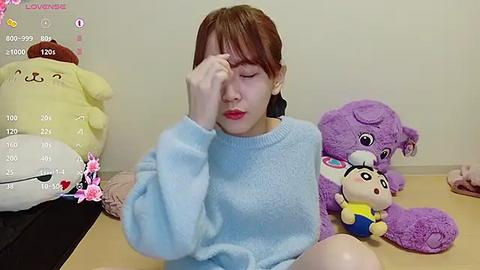 Media: Video of a young Asian woman with reddish-brown hair, wearing a light blue sweater, sitting on a wooden floor. She has a distressed expression with one hand covering her face. Beside her are plush toys including a yellow bear and a purple bear.