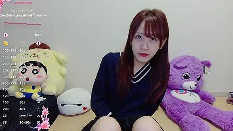 Media: Video of a young Asian woman with long brown hair, wearing a navy sweater, seated on a light-colored surface surrounded by plush toys.