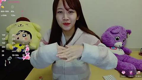 Media: Video of a young Asian woman with long brown hair, wearing a white robe, holding teddy bears with yellow and purple outfits. Background shows a simple room with a wooden desk.