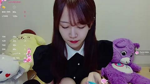 Media: Video of an Asian woman with straight brown hair, red lipstick, black blouse, and red earrings, sitting on a bed surrounded by stuffed animals, including a bear and a yellow duck.