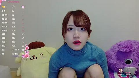 Media: Video of a young Asian woman with red hair, wearing a blue sweater, sitting in front of stuffed animals.