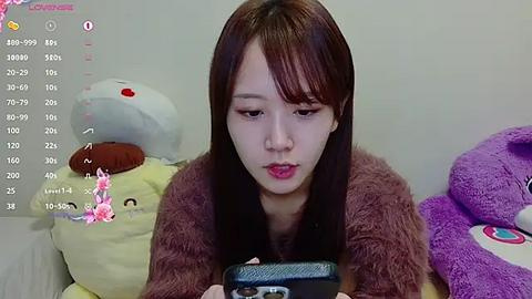 Media: Video of a young East Asian woman with straight, dark brown hair, wearing a brown fur coat, intently using a smartphone, surrounded by plush toys in a cozy bedroom.