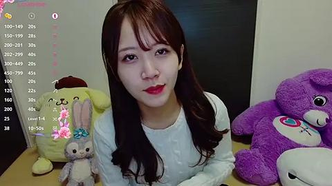 Media: Video of an East Asian woman with long brown hair, wearing a white sweater, sitting among plush toys in a soft, cozy room.