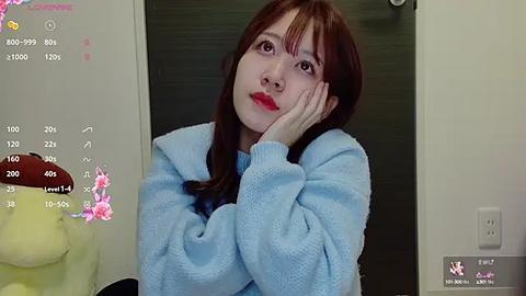 Media: A video of an East Asian woman with long brown hair, wearing a light blue sweater, resting her head on her hands, taken indoors with a calendar and a white wall in the background.