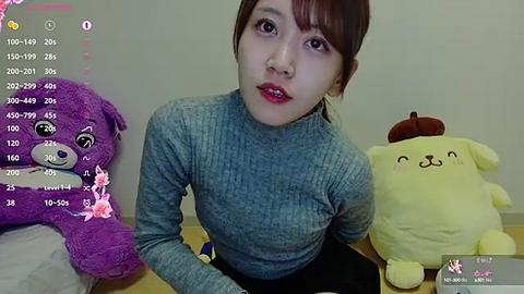 Media: Video of an Asian woman with auburn hair, wearing a grey ribbed turtleneck, sitting on a bed with plush toys: a purple bear, a yellow dog, and a pumpkin.