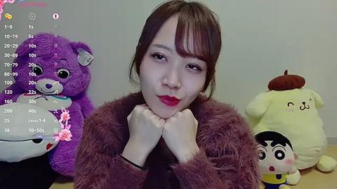 Media: Video of an Asian woman with short brown hair and red lipstick, wearing a brown fur coat, sitting in front of plush toys including a large purple bear, a yellow dog, and a white pig.