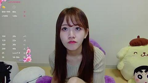 Media: Video of a young East Asian woman with straight brown hair, wearing a beige sweater, sitting in a room with a calendar, plush toys, and pink floral decals on the wall.