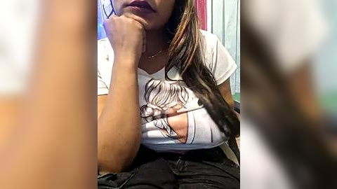 Media: Video of a woman with medium brown skin, long dark hair, and a white graphic tee with a large hole revealing her breast, sitting in a room with a blurred background.