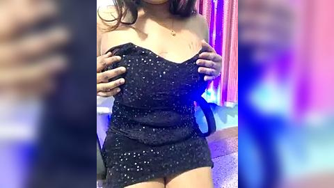 Media: Video of a woman with medium skin tone, wearing a black sequined dress, holding her breasts, indoors with blurred background, pink and blue lighting.