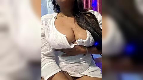 Media: A video of a curvy, dark-skinned woman with long black hair, wearing a white lace bodysuit that accentuates her large breasts, sitting indoors with a blurred background.