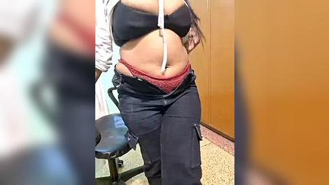 Media: Video of a plus-sized woman with a light brown complexion, wearing a black bra, red panties, and dark pants. She has long hair and is seated on a black examination chair in a medical office with beige walls.