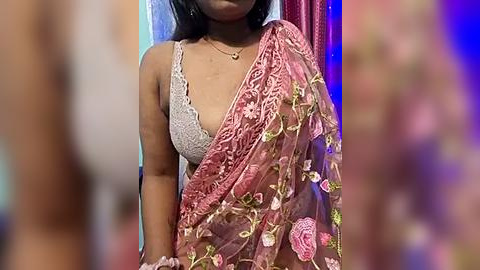 Media: Video of a South Asian woman with medium brown skin, wearing a white lace bra and a pink floral sari, against a blurred background.