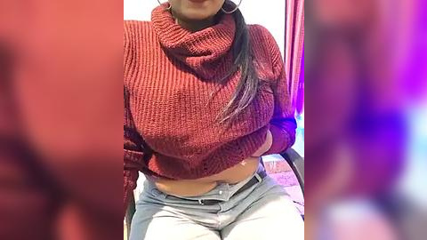 Media: Video of a woman in a red knit sweater and light blue jeans, sitting on a chair with a blurred background.